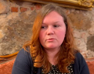 Remigrante Anita Hanna sēž telpā ar akmens sienu fonā