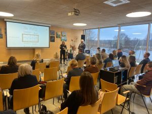 Konferenču zālē notiek prezentācija: pie ekrāna runātāji, zālē sēž klausītāji, fonā lieli logi ar skatu uz dabu