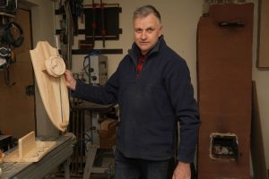 Vīrietis darbnīcā tur rokās paša izgatavotu dekoratīvu koka izstrādājumu; apkārt redzami kokapstrādes instrumenti un iekārtas