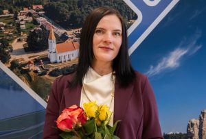 Sieviete formālā apģērbā stāv iekštelpās uz informatīva stenda fona ar ainavām, rokās turot sarkanu un dzeltenu rožu pušķi