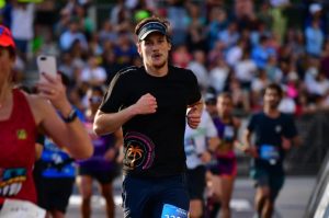 Edgars Prūsis skrien maratonā Maiami, finiša posmā starp citiem skrējējiem, simbolizējot aktīvu dzīvesveidu un atgriešanās ceļu mājās.