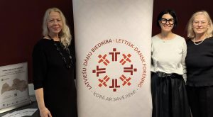 Trīs sievietes stāv pie banera ar uzrakstu “Latviešu–dāņu biedrība” pasākumā Vejlē, Dānijā, kur notika tikšanās ar diasporu par iespējām atgriezties Latvijā