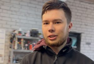 Jauns vīrietis stāv darbnīcā un runā kamerā; fonā redzami instrumenti un plaukti ar dažādiem materiāliem, radot amatniecības un radoša darba vidi