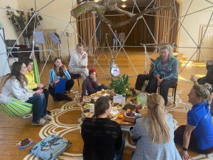 Cilvēku grupa sēž aplī uz koka grīdas gaišā, radošā telpā Augstūjā, Vidzemē, kopīgā pasākumā “Ciemos pie atbraucējiem”