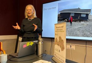 Rīgas plānošanas reģiona remigrācijas koordinatore prezentē remigrācijas tēmu pasākumā telpā ar sarkanu sienu, stāvot pie galda ar datoru, informatīviem materiāliem un brošūrām