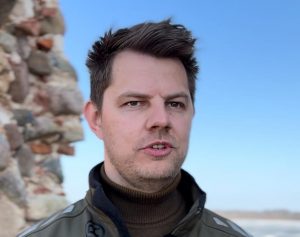 Vīrietis ar īsiem tumšiem matiem stāv ārā pie akmens mūra, fonā redzamas debesis