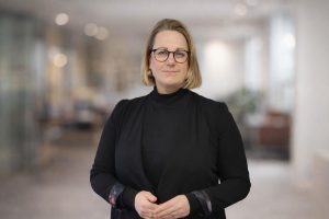 Sieviete ar brillēm un gaišiem, līdz pleciem gariem matiem stāv telpā