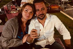 Smaidīgs pāris vakara restorānā. Galds klāts ar ēdieniem un fonā rosās citi viesi.