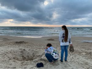 Divi bērni smilšainā pludmalē pie jūras mākoņainā laikā – viens tup un spēlējas ar smiltīm, otrs stāv, rokās turot apavus, skatoties uz viļņiem